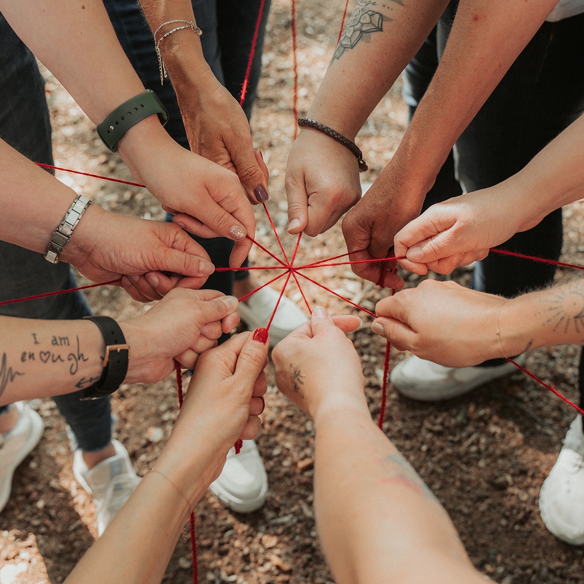 Eine Gruppe von Menschen steht im Kreis, wobei jeder ein rotes Band in der Hand hält, das ihre Hände miteinander verbindet. Das Bild zeigt nur ihre Hände und Unterarme – einige davon mit Tattoos, Armbändern und Uhren – vor einem Hintergrund aus Erde und Blättern.
