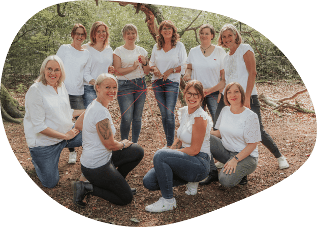 Zehn Frauen, alle in weißen Oberteilen und Jeans gekleidet, posieren gemeinsam in einer waldreichen Umgebung im Freien. Einige knien, und mehrere halten rote Schnüre in den Händen, die sie in einem netzartigen Muster miteinander verbinden – ein visuelles Symbol für Verbundenheit und Chancen, ähnlich wie die Reaktion auf ein Stellenangebot. Alle wirken glücklich und entspannt und spiegeln damit die offene Haltung wider, mit der jede „Apothekerin gesucht“ in ihren Kreis aufgenommen wird.