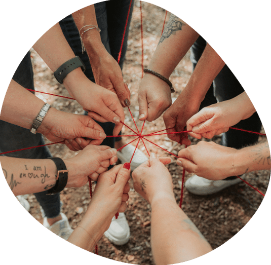 Eine Gruppe von Menschen steht im Kreis und hält rote Schnüre in den Händen, mit denen sie ihre Hände über dem Erdboden miteinander verbinden. Viele von ihnen haben Tätowierungen und Armbänder, was ein Gefühl von Gemeinschaft und Vielfalt vermittelt – vielleicht sogar unter denen, die nach Stellen als Apotheker oder nach einer Stelle als Apothekerin suchen.